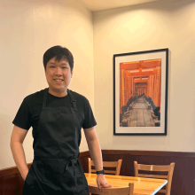 Man in black apron standing in front of a picture inside a Japanese restaurant.